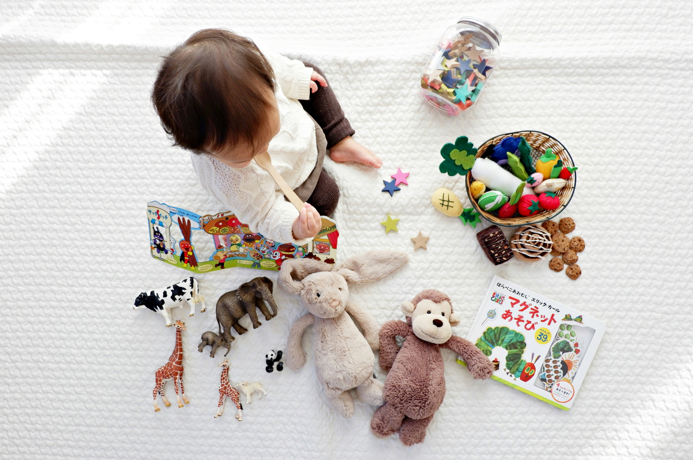 A toddler sitting on a playmat surrounded by soft toys, puzzles and wooden animals