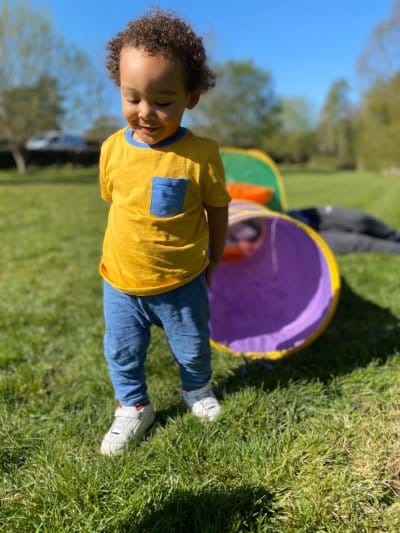 A smiling toddler playing outdoors on the grass beside a colourful pop-up play tunnel