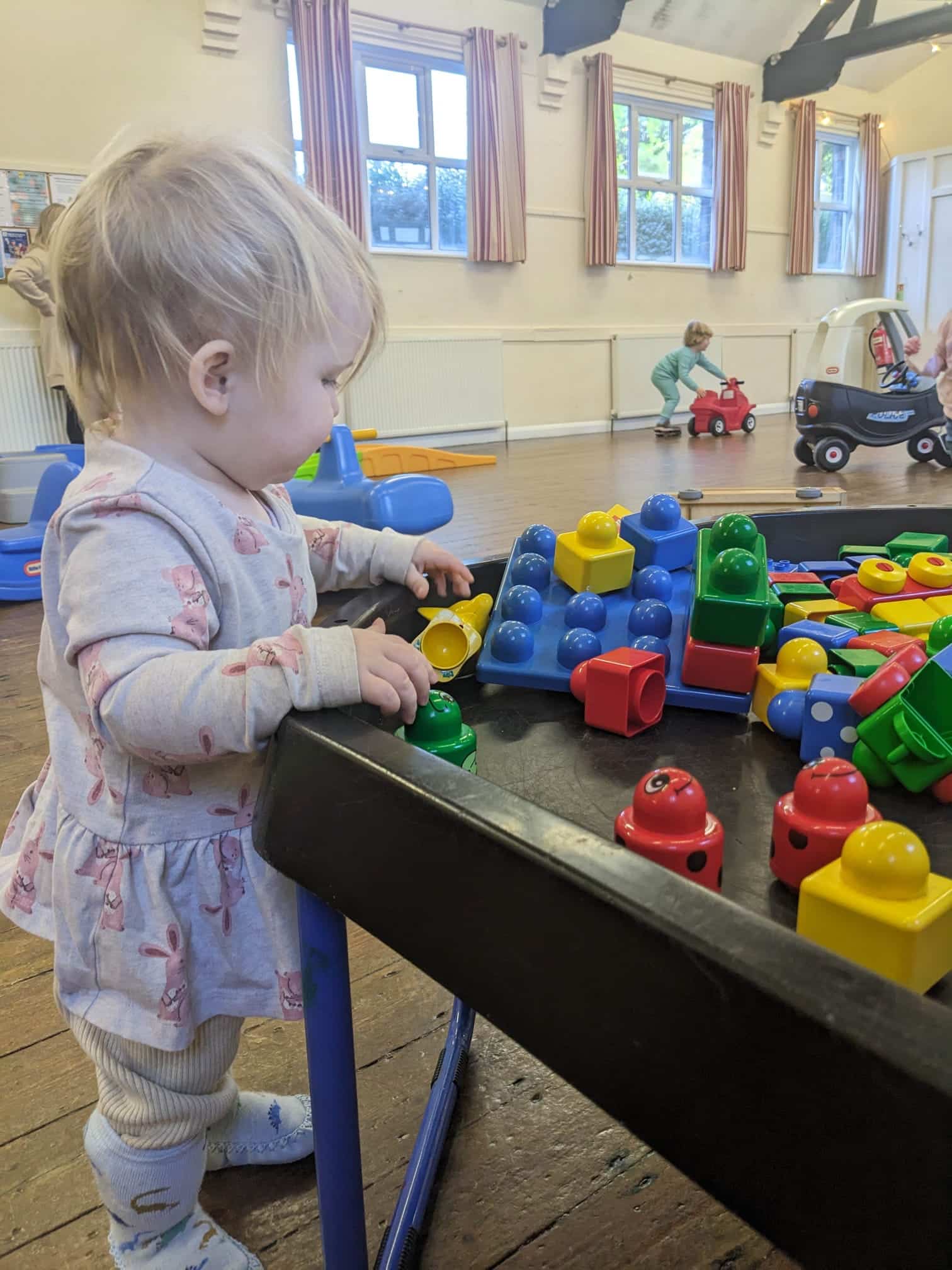 A toddler exploring building blocks at a Stay & Play session while another child rides a toy car behind