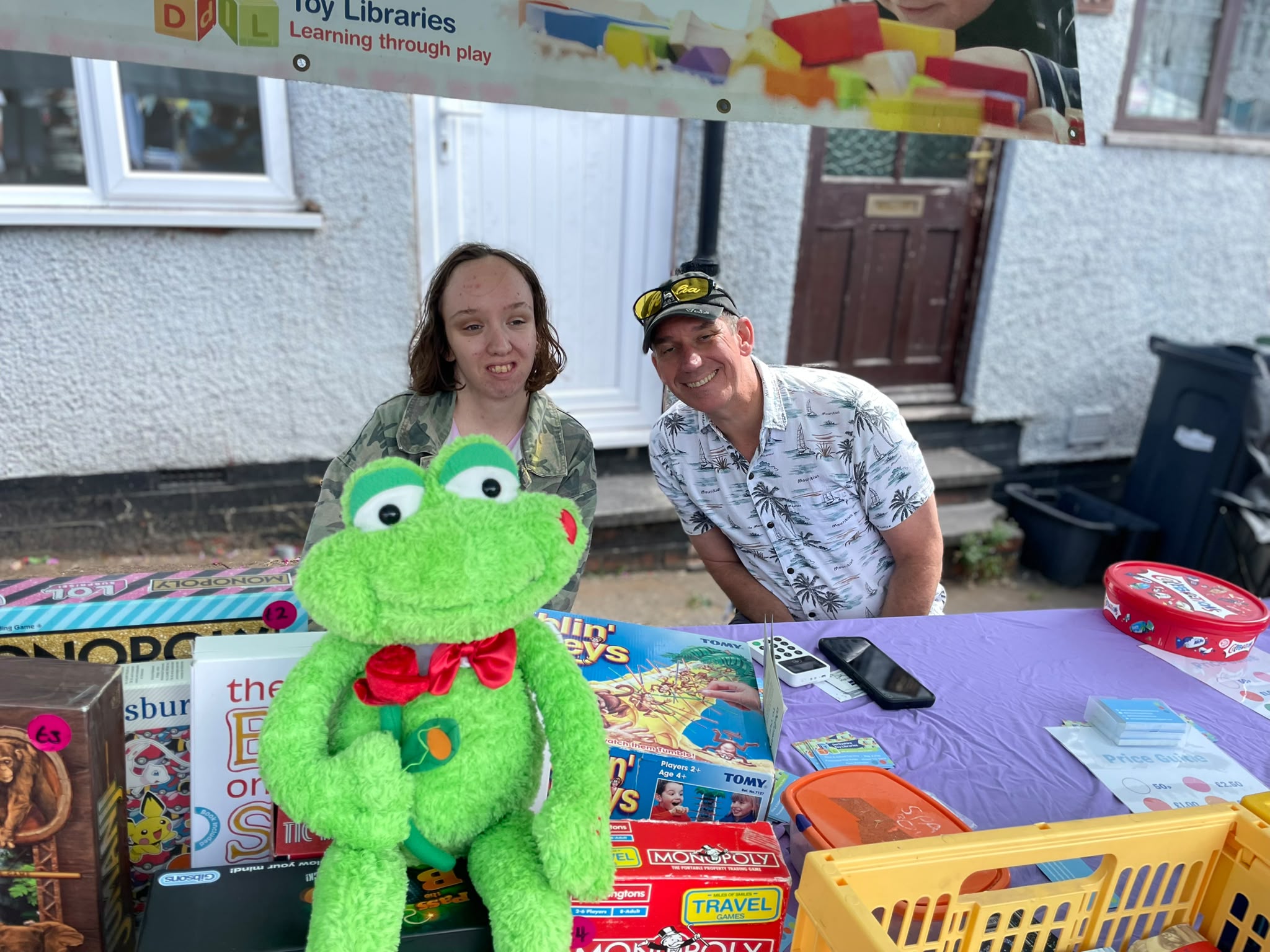 Volunteers with a large green frog plush toy, under a "Learning through play" banner