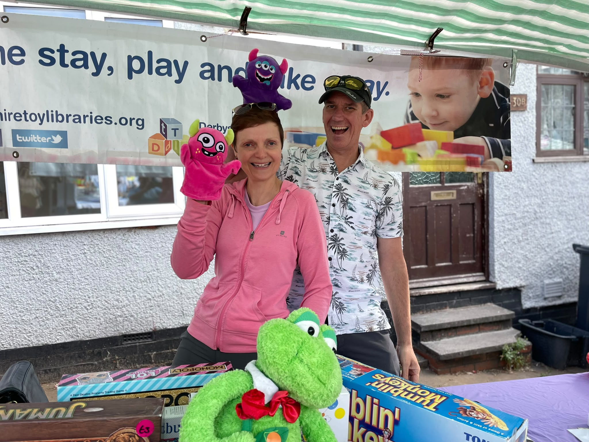 Volunteer wearing pink and purple monster hand puppets, alongside a colleague and the green frog plush