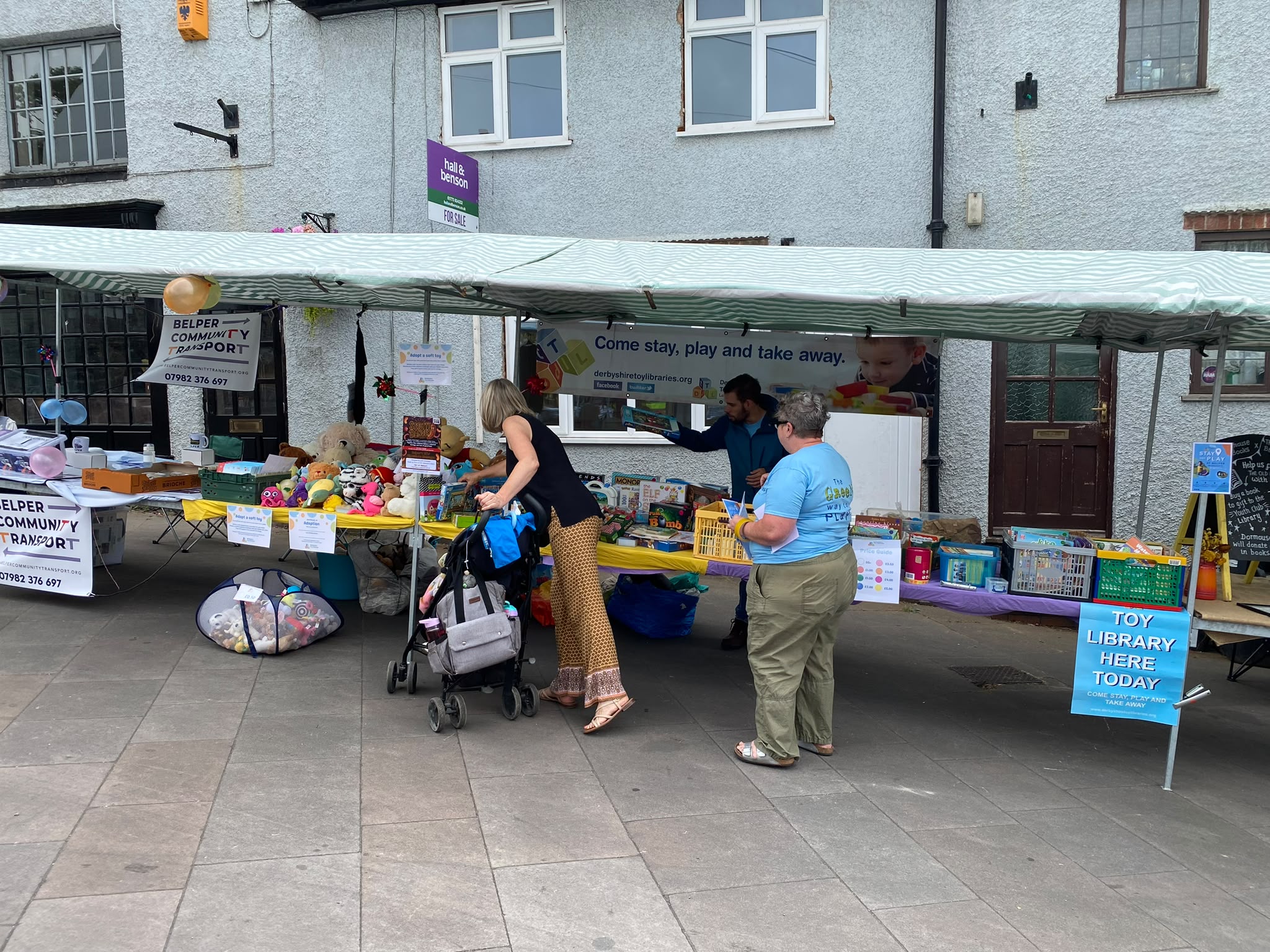 Visitors browsing the toy stall, with a "Toy Library Here Today" sign and a pushchair in shot