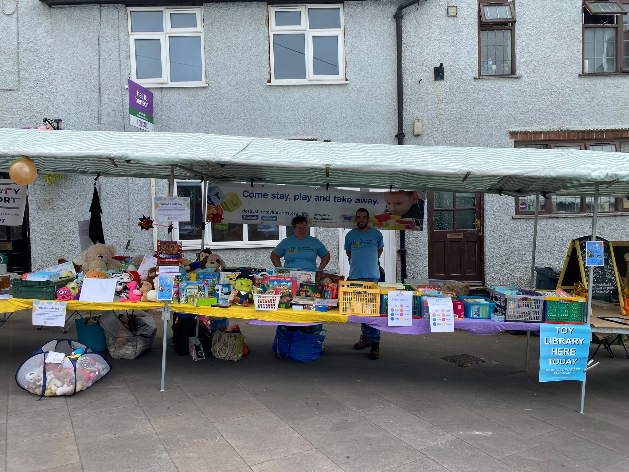 Table laden with soft toys and board games, with an "Adopt a soft toy" sign and a "Toy Library Here Today" sign nearby