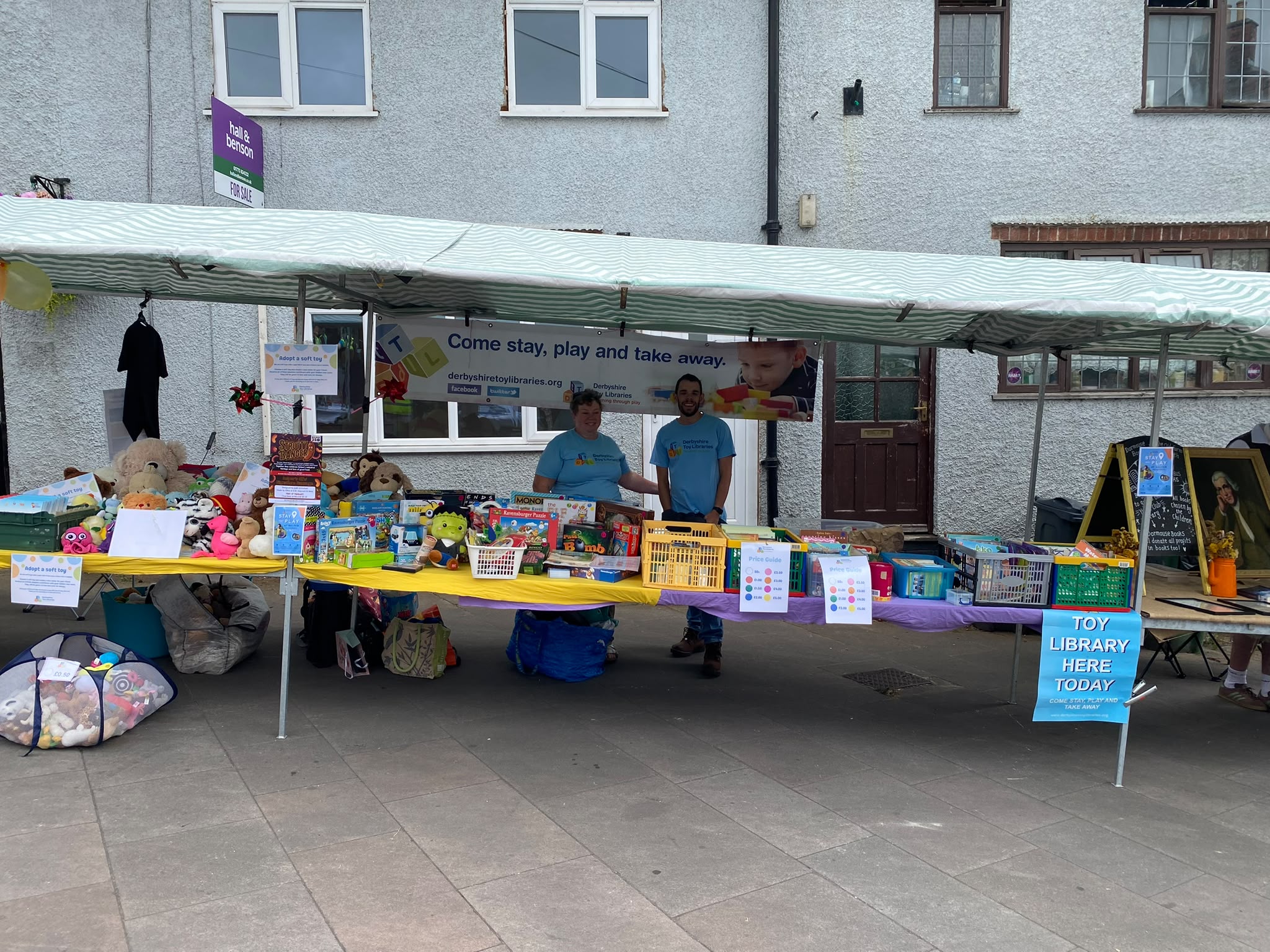 Two volunteers behind the toy and game table, with soft toys and the "Toy Library Here Today" sign